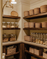 A cozy traditional pantry featuring warm cream walls, rich dark wood shelving, and a mix of woven rattan baskets and clear glass jars holding pantry staples. An aged brass semi-flush mount light fixture illuminates the space.