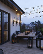 Industrial deck at dusk featuring warm white Edison bulb string lights draped overhead, dark bronze gooseneck wall sconces, a raw wood dining table, and large black metal lanterns with LED candles. — alternate angle