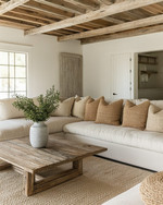 A cozy farmhouse poolhouse interior featuring a large cream sectional sofa with woven throw pillows, a distressed wood coffee table, and an exposed reclaimed wood ceiling. Soft light filters in, highlighting a natural fiber rug and a simple ceramic vase with greenery. — styling and décor detail