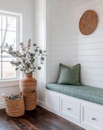 A bright farmhouse mudroom with white shiplap walls, a built-in bench with a sage green cushion, and dark wood flooring. Seasonal touches include a vase of eucalyptus, woven baskets, and a terracotta pot with dried botanicals, all bathed in warm, natural light. — alternate angle