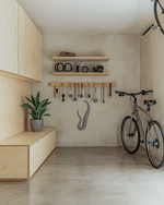 A small Japandi garage featuring light birch plywood cabinetry, a polished concrete floor, and walls painted in a soft off-white. A minimalist bamboo storage bench sits against one wall, with a single snake plant in a grey ceramic pot. Wall-mounted racks hold neatly organized tools, and a bicycle is suspended, maximizing floor space.