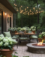 A cozy traditional patio featuring a wrought iron dining set with sage green cushions, a wooden dining table, and warm string lights draped overhead. Terracotta pots filled with lush greenery and blooming hydrangeas are placed around a stone fire pit.