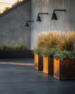 An industrial driveway featuring dark gray sealed concrete, bordered by robust steel edging, with matte black gooseneck lighting fixtures mounted on a raw concrete wall and large Corten steel planters filled with hardy ornamental grasses. — close-up of textures and materials