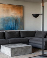 A modern family room featuring a large charcoal grey sectional sofa, a sculptural concrete coffee table, and a large abstract painting with blues and sienna tones. A tall, slender black metal floor lamp stands next to the sofa. — close-up of textures and materials