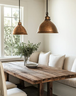 A warm and inviting farmhouse breakfast nook featuring a rustic reclaimed wood table, a cream banquette with linen cushions, and two aged bronze pendant lights hanging over the table, with soft morning light streaming through a window. — alternate angle