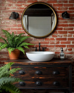 Industrial bathroom with a warm red exposed brick wall, a dark wood reclaimed vanity with a white ceramic basin, and matte black fixtures. A large round mirror with an aged brass frame hangs above the vanity, complemented by a lush green fern in a terracotta pot. — alternate angle