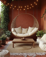 A cozy boho porch featuring a rattan hanging chair, a jute rug, and a low wooden coffee table surrounded by plush cream cushions. Fairy lights are strung overhead, and various potted plants add greenery against a backdrop of warm terracotta walls. — alternate angle