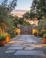 Rustic driveway with warm grey gravel and large, irregular flagstones leading to a wooden gate. The scene is enhanced by low-level pathway lights, subtle uplighting on surrounding trees, and terracotta planters filled with native greenery, all under a soft, twilight glow. — close-up of textures and materials