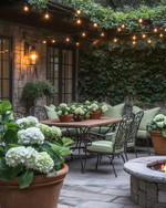 A cozy traditional patio featuring a wrought iron dining set with sage green cushions, a wooden dining table, and warm string lights draped overhead. Terracotta pots filled with lush greenery and blooming hydrangeas are placed around a stone fire pit. — close-up of textures and materials