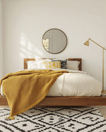 A renter-friendly Mid-Century Modern bedroom featuring a walnut platform bed with a cream duvet, a mustard yellow knit throw, and a graphic black and white area rug. Brass multi-arm floor lamp and a simple round mirror accent the space. — alternate angle
