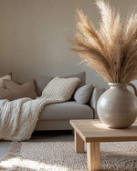 A cozy modern landscape living room featuring a warm gray sofa, a chunky cream knit throw, a light oak coffee table, and a large ceramic vase with dried pampas grass, all illuminated by soft natural light. — close-up of textures and materials