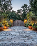 Rustic driveway with warm grey gravel and large, irregular flagstones leading to a wooden gate. The scene is enhanced by low-level pathway lights, subtle uplighting on surrounding trees, and terracotta planters filled with native greenery, all under a soft, twilight glow.