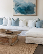 A serene coastal family room featuring a large linen slipcovered sectional sofa, a reclaimed wood coffee table, and a woven jute rug. Soft blue and white throw pillows add subtle color, complemented by a large abstract ocean-inspired artwork above the sofa and natural rattan accents.
