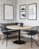 A modern breakfast nook features a light wood round table with a minimalist black metal base, paired with a charcoal grey upholstered bench and two black metal chairs. A subtle geometric peel-and-stick wallpaper adorns the wall, alongside a gallery of abstract art prints. — close-up of textures and materials