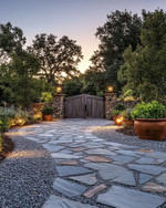 Rustic driveway with warm grey gravel and large, irregular flagstones leading to a wooden gate. The scene is enhanced by low-level pathway lights, subtle uplighting on surrounding trees, and terracotta planters filled with native greenery, all under a soft, twilight glow. — styling and décor detail