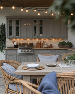 Cozy coastal outdoor kitchen featuring weathered gray cabinetry, honed travertine countertops, a teak dining table with comfortable cushioned chairs, and warm string lights overhead, accented with woven rattan and soft indigo textiles. — close-up of textures and materials