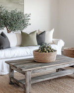 A cozy farmhouse living room featuring a cream slipcovered sofa, a distressed wooden coffee table, and a jute area rug. Neutral tones dominate, with accents of charcoal and muted green in throw pillows and a large olive tree in a woven basket, all bathed in soft, inviting light. — close-up of textures and materials