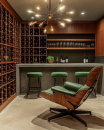 Mid-Century Modern wine cellar featuring a walnut Eames Lounge Chair, a Sputnik chandelier, sleek teak wine racks, and a concrete tasting bar with green upholstered stools. — alternate angle
