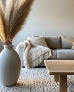 A cozy modern landscape living room featuring a warm gray sofa, a chunky cream knit throw, a light oak coffee table, and a large ceramic vase with dried pampas grass, all illuminated by soft natural light. — alternate angle