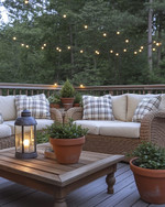 A cozy farmhouse deck features deep-seated wicker furniture with cream cushions and plaid throw pillows, illuminated by warm string lights and a metal lantern. Terracotta pots with lush greenery and a rustic wooden coffee table complete the inviting outdoor scene. — close-up of textures and materials