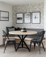 A modern breakfast nook features a light wood round table with a minimalist black metal base, paired with a charcoal grey upholstered bench and two black metal chairs. A subtle geometric peel-and-stick wallpaper adorns the wall, alongside a gallery of abstract art prints.