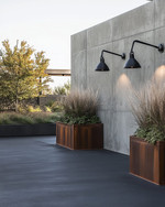 An industrial driveway featuring dark gray sealed concrete, bordered by robust steel edging, with matte black gooseneck lighting fixtures mounted on a raw concrete wall and large Corten steel planters filled with hardy ornamental grasses. — alternate angle