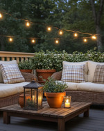 A cozy farmhouse deck features deep-seated wicker furniture with cream cushions and plaid throw pillows, illuminated by warm string lights and a metal lantern. Terracotta pots with lush greenery and a rustic wooden coffee table complete the inviting outdoor scene. — styling and décor detail