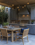 Cozy coastal outdoor kitchen featuring weathered gray cabinetry, honed travertine countertops, a teak dining table with comfortable cushioned chairs, and warm string lights overhead, accented with woven rattan and soft indigo textiles. — alternate angle