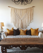 A Farmhouse Craft living room featuring a large, hand-carved reclaimed oak coffee table, a woven macrame wall hanging in ivory, and a wrought-iron chandelier, with ceramic table lamps and deep ochre throw pillows. — styling and décor detail