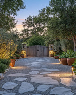 Rustic driveway with warm grey gravel and large, irregular flagstones leading to a wooden gate. The scene is enhanced by low-level pathway lights, subtle uplighting on surrounding trees, and terracotta planters filled with native greenery, all under a soft, twilight glow. — alternate angle