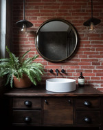 Industrial bathroom with a warm red exposed brick wall, a dark wood reclaimed vanity with a white ceramic basin, and matte black fixtures. A large round mirror with an aged brass frame hangs above the vanity, complemented by a lush green fern in a terracotta pot.