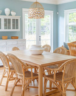 A coastal dining room featuring a light oak modern dining table, vintage rattan chairs, a distressed white buffet, and a contemporary capiz shell chandelier, with walls painted in a soft Palladian Blue. — alternate angle