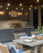 Cozy coastal outdoor kitchen featuring weathered gray cabinetry, honed travertine countertops, a teak dining table with comfortable cushioned chairs, and warm string lights overhead, accented with woven rattan and soft indigo textiles. — styling and décor detail