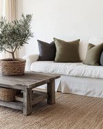 A cozy farmhouse living room featuring a cream slipcovered sofa, a distressed wooden coffee table, and a jute area rug. Neutral tones dominate, with accents of charcoal and muted green in throw pillows and a large olive tree in a woven basket, all bathed in soft, inviting light. — alternate angle