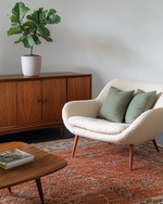 A Mid-Century Modern living room featuring a rich walnut credenza, a teak coffee table, and a cream bouclé lounge chair. The space is anchored by a wool rug in deep terracotta, accented with sage green linen pillows, and houses a tall fiddle-leaf fig in an unglazed ceramic planter. — styling and décor detail