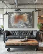 An industrial landscape living room featuring a charcoal distressed leather Chesterfield sofa, a reclaimed wood coffee table on caster wheels, and a large abstract painting in grey and burnt sienna tones, under exposed metal pendant lighting. — alternate angle