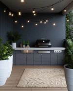 A modern outdoor kitchen setup featuring a charcoal gray modular grill station with a light concrete-effect countertop, brushed stainless steel accents, and black string lights strung overhead. Large white ceramic planters with architectural green plants frame the area, complemented by a woven outdoor rug in a subtle geometric pattern. — alternate angle