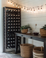 A cozy farmhouse wine cellar featuring a dark stained wooden wine rack filled with bottles, a rustic console table with a decanter, and warm string lights with Edison bulbs draped across a temporary shiplap wall in antique white. A few woven baskets and ceramic planters with subtle greenery complete the inviting, rustic aesthetic. — close-up of textures and materials