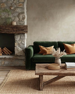 A Rustic Landscape living room featuring a deep forest green velvet sofa, a reclaimed wood coffee table, and a rough-hewn stone fireplace. Natural light illuminates the warm greige walls, complemented by woven jute rugs and subtle ochre throw pillows.