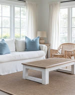 A serene coastal sunroom featuring a slipcovered sofa in a creamy off-white, paired with rattan chairs and a bleached oak coffee table. Soft dusty blue throw pillows and a woven jute rug add subtle color accents, all illuminated by ample natural light filtering through sheer linen curtains.