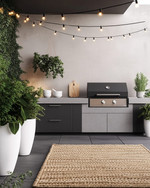 A modern outdoor kitchen setup featuring a charcoal gray modular grill station with a light concrete-effect countertop, brushed stainless steel accents, and black string lights strung overhead. Large white ceramic planters with architectural green plants frame the area, complemented by a woven outdoor rug in a subtle geometric pattern. — close-up of textures and materials
