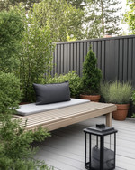 A Scandinavian backyard featuring a light grey wooden deck, a simple slatted teak bench with a charcoal grey outdoor cushion, and a minimalist black metal lantern. Greenery includes potted evergreen shrubs and ornamental grasses in terracotta pots, creating a serene, natural ambiance. — styling and décor detail