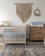Coastal baby and kids' room with a white oak crib, a large seagrass rug, a rattan changing table, and a macrame wall hanging above a dresser. Soft white walls are accented with muted blue textiles and natural wood elements. — styling and décor detail