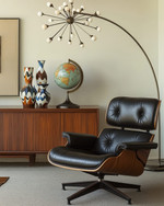 A sophisticated Mid-Century Modern library featuring an Eames Lounge Chair in black leather and walnut, an Arco floor lamp arching over, and a teak credenza displaying abstract ceramic vases and a vintage globe, all under the warm glow of a Sputnik chandelier. — alternate angle
