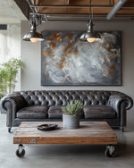 An industrial landscape living room featuring a charcoal distressed leather Chesterfield sofa, a reclaimed wood coffee table on caster wheels, and a large abstract painting in grey and burnt sienna tones, under exposed metal pendant lighting. — styling and décor detail