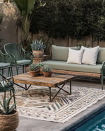 A boho poolside area featuring a mix of vintage wrought-iron chairs in sage green and a modern low-slung outdoor sofa with natural linen cushions. A reclaimed wooden coffee table sits on a layered outdoor rug with geometric patterns, surrounded by potted succulents and woven rattan accents.