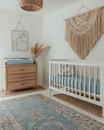 Coastal baby and kids' room with a white oak crib, a large seagrass rug, a rattan changing table, and a macrame wall hanging above a dresser. Soft white walls are accented with muted blue textiles and natural wood elements.