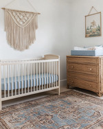 Coastal baby and kids' room with a white oak crib, a large seagrass rug, a rattan changing table, and a macrame wall hanging above a dresser. Soft white walls are accented with muted blue textiles and natural wood elements. — alternate angle