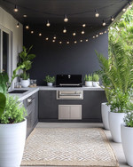 A modern outdoor kitchen setup featuring a charcoal gray modular grill station with a light concrete-effect countertop, brushed stainless steel accents, and black string lights strung overhead. Large white ceramic planters with architectural green plants frame the area, complemented by a woven outdoor rug in a subtle geometric pattern. — styling and décor detail