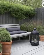 A Scandinavian backyard featuring a light grey wooden deck, a simple slatted teak bench with a charcoal grey outdoor cushion, and a minimalist black metal lantern. Greenery includes potted evergreen shrubs and ornamental grasses in terracotta pots, creating a serene, natural ambiance. — alternate angle